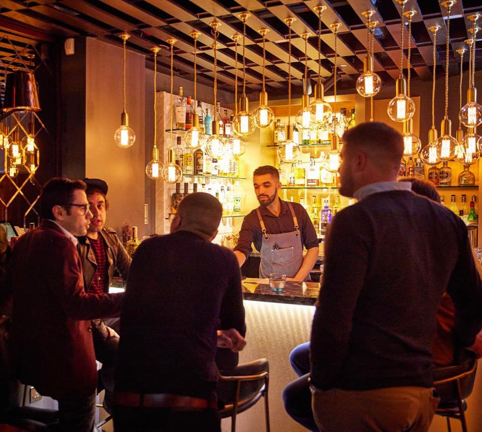 A warmly lit London bar with pendant Edison bulbs, a bartender in an apron behind the counter, and customers at the bar mid-service.